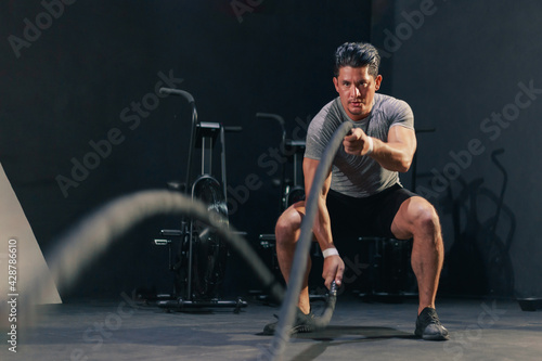 Young fit handsome muscular man in activewear working out heavy battle rope at the gym. Motivation concentration endurance exercise