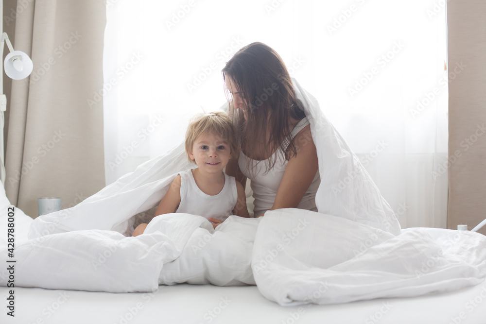 Beautiful blond child and his mother, playing at home in the morning in bed, smiling, laughing and having fun Beautiful blond child and his mother, playing at home in the morning in bed, smiling, laughing and having fun