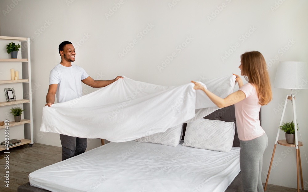 Fototapeta premium Cheerful multiracial couple making bed in morning, having fun at home
