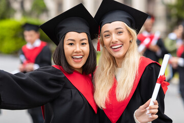 Happy multiracial young ladies in graduation clothes taking selfie
