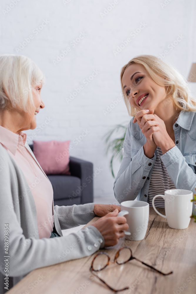 Positive woman talking to mother with cup of tea near eyeglasses Positive woman talking to mother with cup of tea near eyeglasses