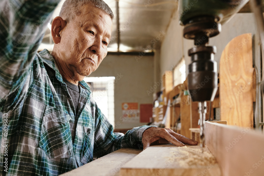 Elderly Asian carpenter using drilling machine to cut clean holes in wooden board