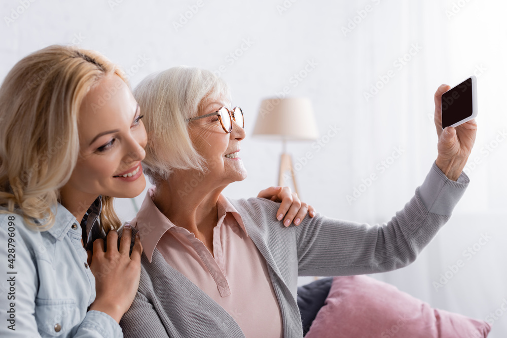 Smiling elderly parent taking selfie with cellphone near daughter Smiling elderly parent taking selfie with cellphone near daughter