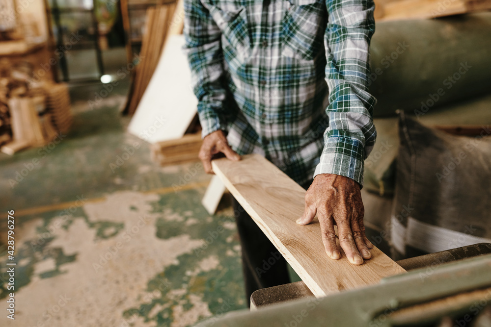 Hands of aged carpenter putting wooden board in thickness planer on workbench