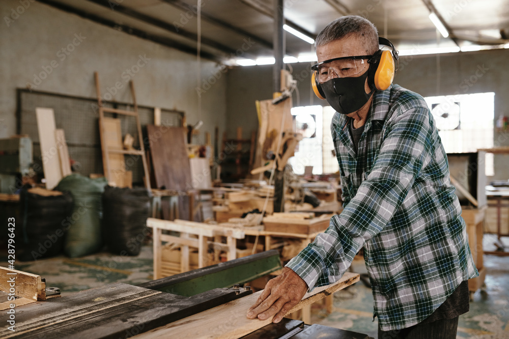 Senior man in protective mask and earmuffs working with wood in carpenters workshop Senior man in protective mask and earmuffs working with wood in carpenters workshop