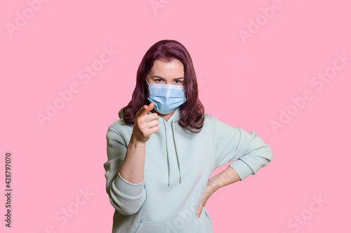 young woman in a medical mask and green hoodie points a finger to the camera. Virus protection and security concept. Isolated on pink