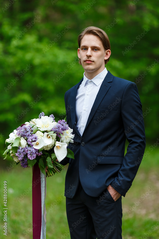 Young beautiful groom in dark blue suit standing and holding big wedding bouquet with lilac and long ribbons. Spring, park outdoor Young beautiful groom in dark blue suit standing and holding big wedding bouquet with lilac and long ribbons. Spring, park outdoor