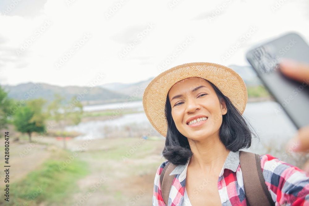 woman stand on the banks of the lake using mobile phone takes a picture selfie, taking pictures, Video call, Asian woman hiker in front smiling happy, Woman hiking in woods, warm summer day.