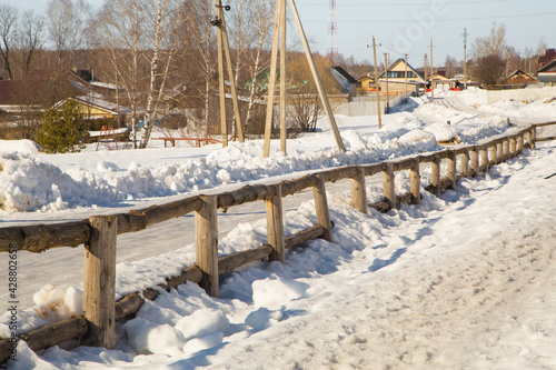 Beautiful countryside in winter with wooden fence and houses in the distance. Wonderful rural landscape with wooden houses and a snow-covered road behind a hedge.