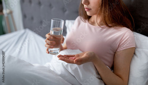 Cropped view of young woman with glass of water taking sleeping pills in bed, copy space