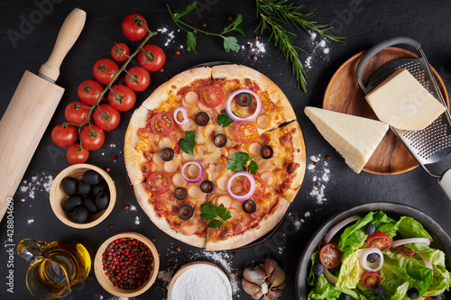 assorted foodset on table. Italian pizza and pizza cooking ingredients on dark stone background. tomatoes on vine, mozzarella, black olives, herbs and spices, vegetable salad. top view. photo