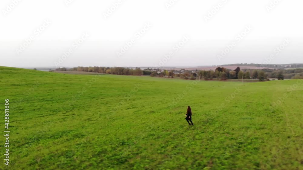 Shooting from drone of a young girl in a black trench coat walking across a huge green field against the backdrop sunset Shooting from drone of a young girl in a black trench coat walking across a huge green field against the backdrop sunset