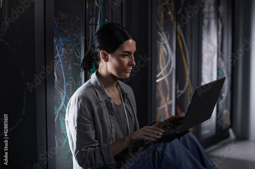 Side view portrait of female IT engineer using laptop in server room while working with supercomputer at data center, copy space