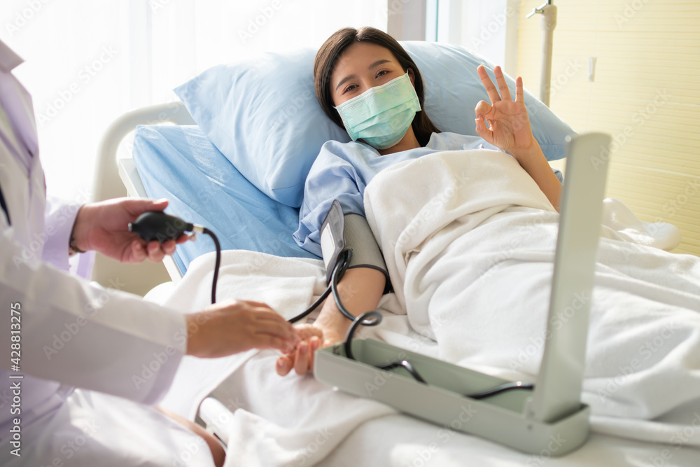 Happy Asian female patient wears a mask, lies on the bed, and Shows an ok symbol. When the doctor examines the symptoms using a pressure gauge. Concept of believe in treatment And insurance coverage Happy Asian female patient wears a mask, lies on the bed, and Shows an ok symbol. When the doctor examines the symptoms using a pressure gauge. Concept of believe in treatment And insurance coverage