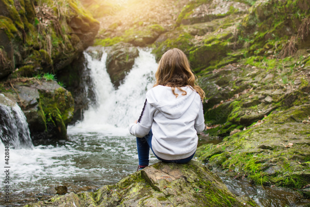 Young girl sitting on the rock and looking at waterfall on the river in the rocks. Tourism with children concept.