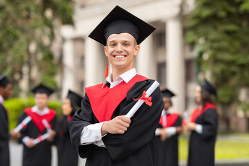 Cheerful guy in graduation costume with diploma posing outdoors