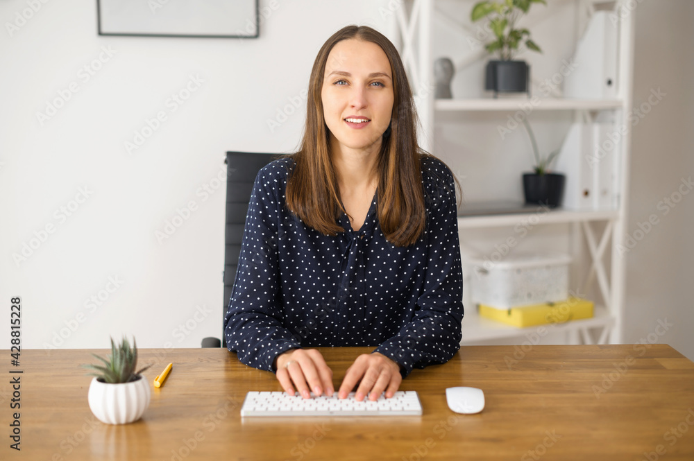 Modern young business woman is typing on the keyboard and looks at the camera. Webcam view at the female office employee answering emails, messaging online, programming Modern young business woman is typing on the keyboard and looks at the camera. Webcam view at the female office employee answering emails, messaging online, programming