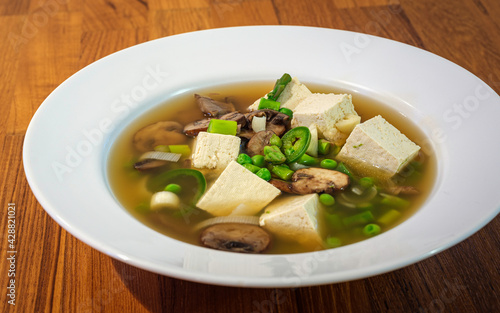 Tofu Mushroom Soup with Green Peas and Onion in Vegetable Broth. Healthy Vegetarian Soup in a White flat Bowl on Brown Teakwood Table. 