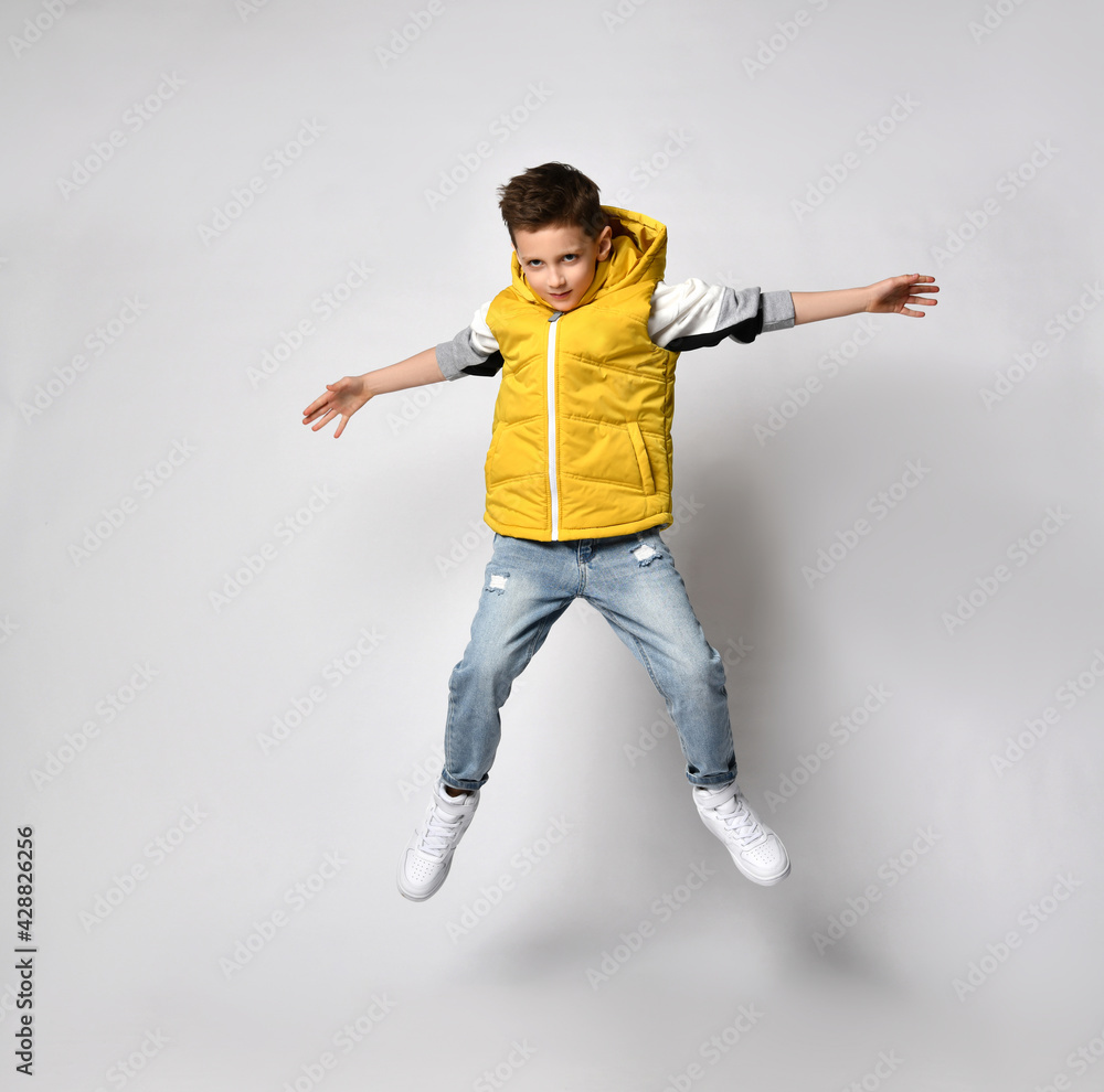 teenage guy in a yellow puffed vest, a sleeveless down jacket with a hood and jeans is having fun and jumping high on a white background