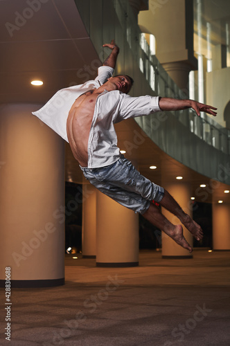 Young male ballet dancer, wearing in white shirt and jeans, jumping on a building background. Outdoor dance.