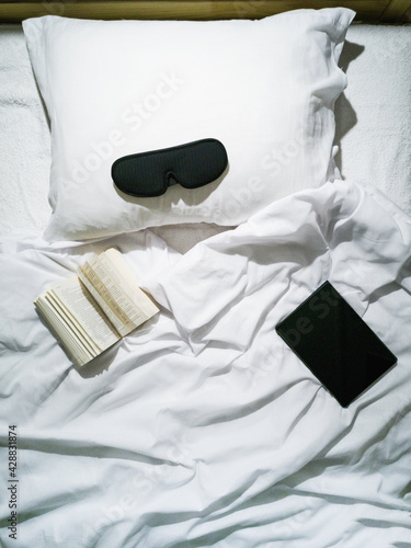 On the bed lies a tablet, an open book, and a sleep mask