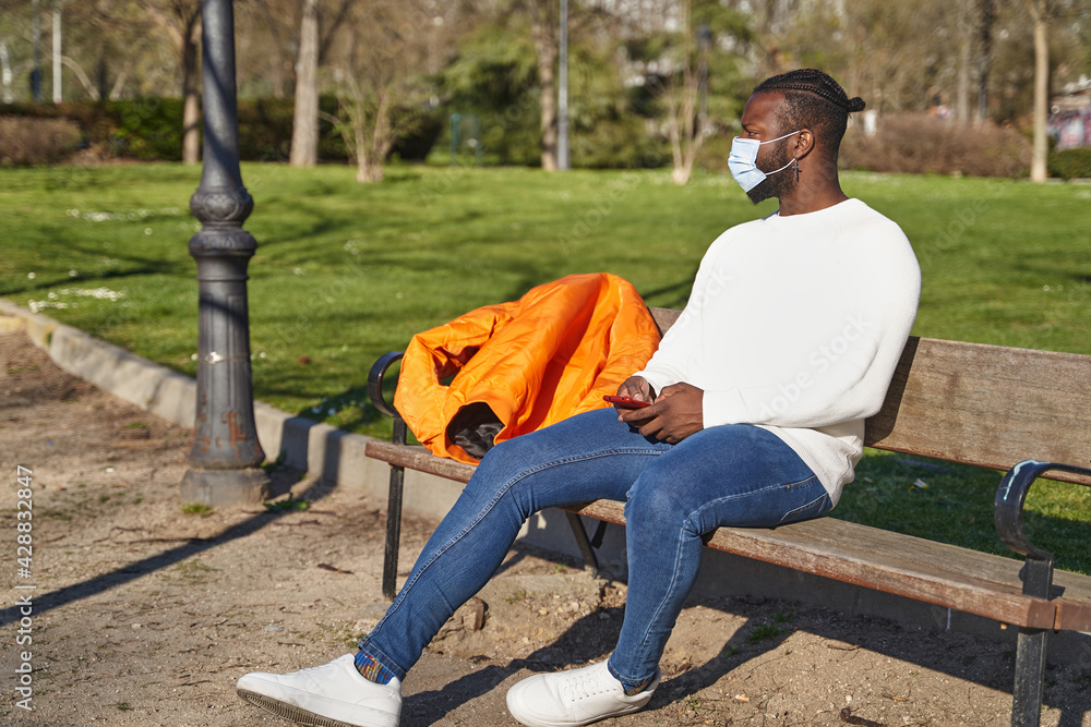 Young African American man sitting on a park bench sending a message. Latino man with anti-contagion mask. Concept of new normal. Young African American man sitting on a park bench sending a message. Latino man with anti-contagion mask. Concept of new normal.