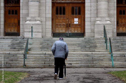 Montreal, Quebec, Canada - 17 April 2021: during Covid-19 coronavirus pandemic crisis, a lonely old man with grey hair walks towards a closed church, looking for peace in religion.