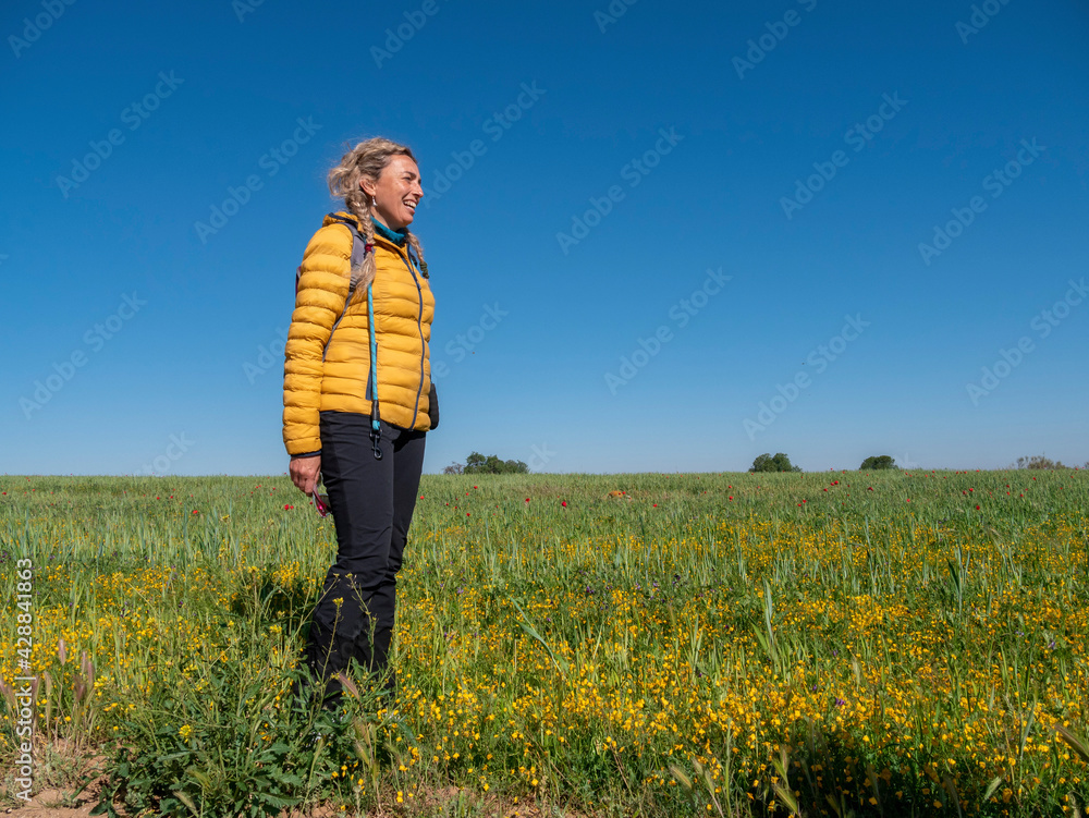 ATTRACTIVE BLONDE WOMAN WITH HIKING CLOTHES AND BACKPACK, POSING ON A SUNNY SPRING DAY IN THE GREEN FIELDS OF TOLEDO, SPAIN