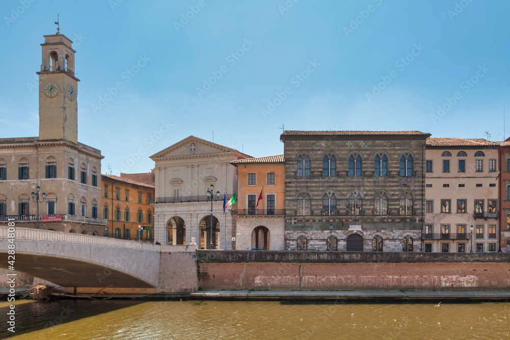 The Palazzo Pretorio and its clock tower in front of the Ponte Di Mezzo and next to the Logge Dei Banchi, the city hall and Palazzo Gambacorti alongside the Arno River in Pisa, Italy