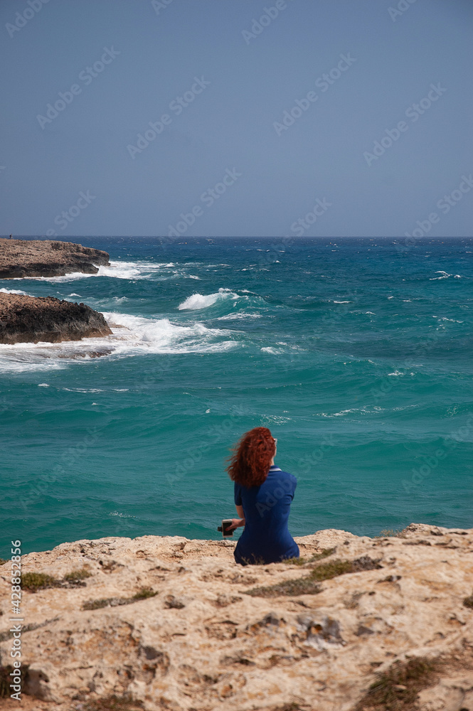 A girl in a dress sits alone on the rocky shore of the turquoise blue sea