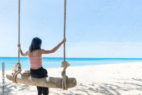 Back Beautiful young Asian women with tan skin wearing pink tank black pants sitting on timber swing. beach and seaside fresh sky background. Holiday, Vacation, Chilling, Relax concept. Alone Concept.