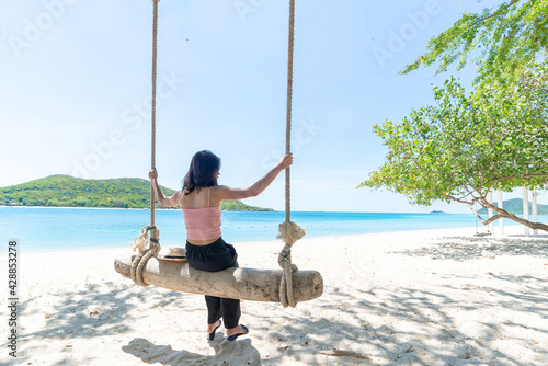 Back Beautiful young Asian women with tan skin wearing pink tank black pants sitting on timber swing. beach and seaside fresh sky background. Holiday, Vacation, Chilling, Relax concept. Alone Concept.
