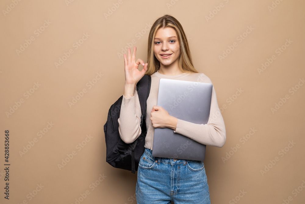 Portrait of young woman holding closed laptop with positive friendly smile standing isolated on beige background Portrait of young woman holding closed laptop with positive friendly smile standing isolated on beige background