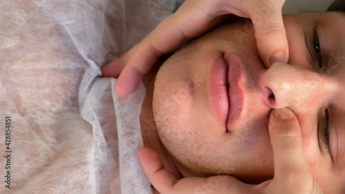 Manual therapist doctor is making facial massage for young man, closeup face top view. Treatment of headaches with physiotherapist. She presses her fingers on certain points on the patient's face.