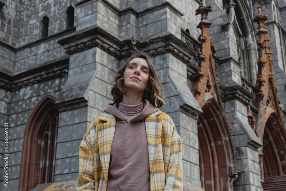 Naklejka premium Happy millenial woman with closed eyes enjoys in a yellow coat in European city near historic building. Local travel concept.