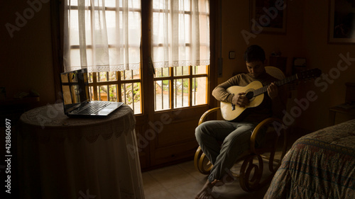 Learning guitar at home with laptop. Happy young man playing guitar while sitting on the sofa at home.