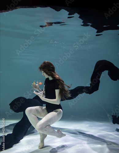 beautiful girl in a black swimsuit with a black pareo plays with fish in an aquarium on a blue background underwater