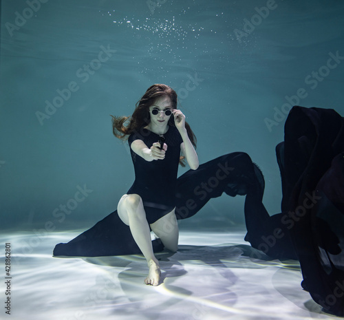 beautiful girl in a black swimsuit with a black pareo holds a black pistol in her hands on a blue background underwater © константин константи