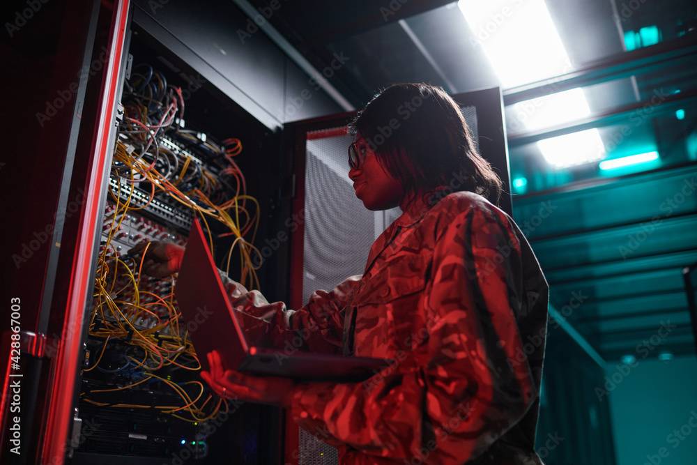 Low angle portrait of African-American woman managing computer network while working with supercomputer in server room lit by red light, copy space