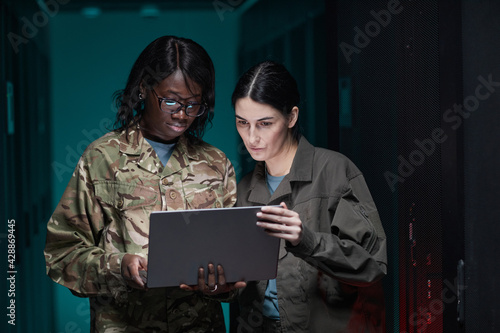 Waist up portrait of two young women wearing military uniform using laptop while standing in server room, copy space
