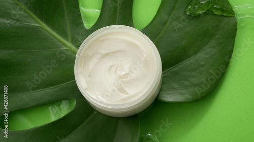 Cosmetic jar with cream on green tropical leaf in clean water splash on green background. Top view. Plant-based skincare, organic natural ingredients, skin routines, summer cosmetics, fresh concept
