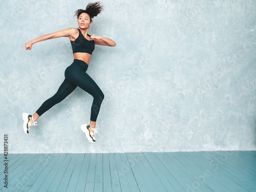 Fitness smiling black woman in sports clothing with afro curls hairstyle.She wearing sportswear. Young beautiful model with perfect tanned body.Female jumping and running in studio near gray wall