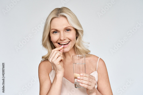 Happy middle aged 50s woman holding pill and glass of water taking dietary supplements. Portrait of smiling adult attractive woman taking care of health in menopause, isolated on white. photo