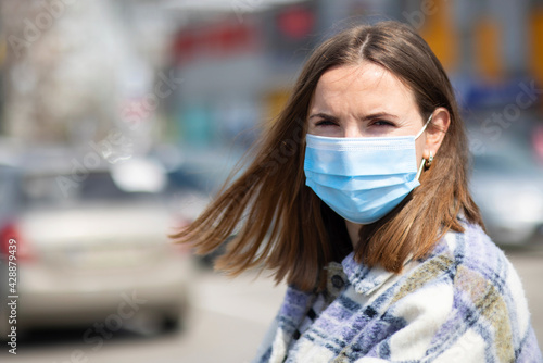 A young woman in a medical mask stands in a parking lot near a grocery store. A woman is protecting herself from the coronavirus. Shopping during quarantine
