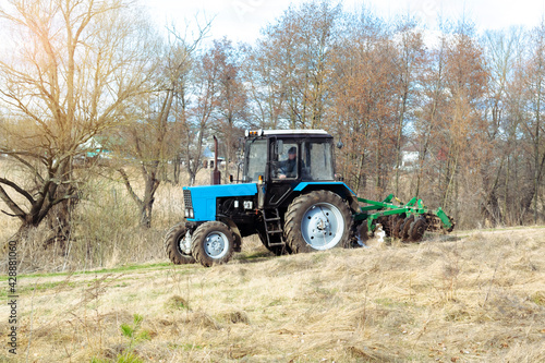 Blue small modern tractor machinery plowing agricultural field meadow at farm on sunny day. Farmer cultivating and make soil tillage before seeding plants and crops  nature countryside rural scene.