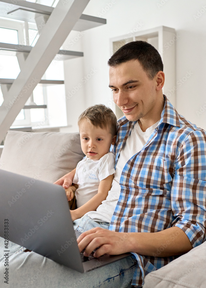 Happy young adult dad embracing cute small kid son looking at laptop device sitting on couch. Smiling father teaching toddler child using computer at home learning online, playing, watching cartoons. Happy young adult dad embracing cute small kid son looking at laptop device sitting on couch. Smiling father teaching toddler child using computer at home learning online, playing, watching cartoons.