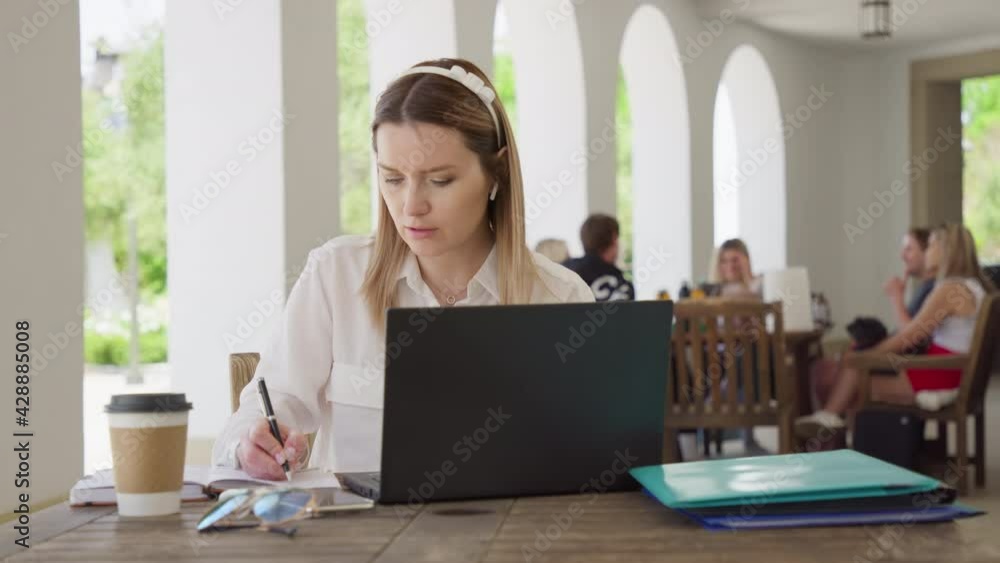 Successful young beautiful blonde woman with laptop and coffee cup sitting outdoors in modern business center park and making notes in planner. Career people, focused business woman. RED footage 8K