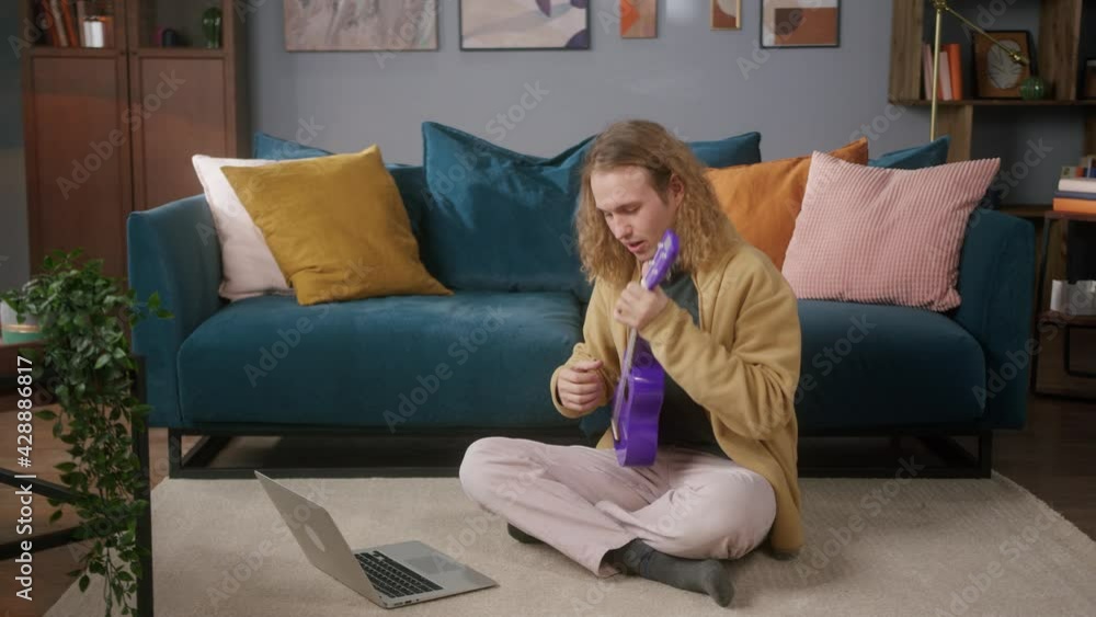 Happy man using laptop computer playing purple ukulele sitting on floor. Blogger Teacher teaching how to play on music instrument online. Remote musical learning. 