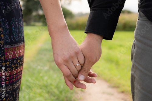couple holding hands, close up she has an engagement ring on her finger in the foreground with an unfocused nature background.
