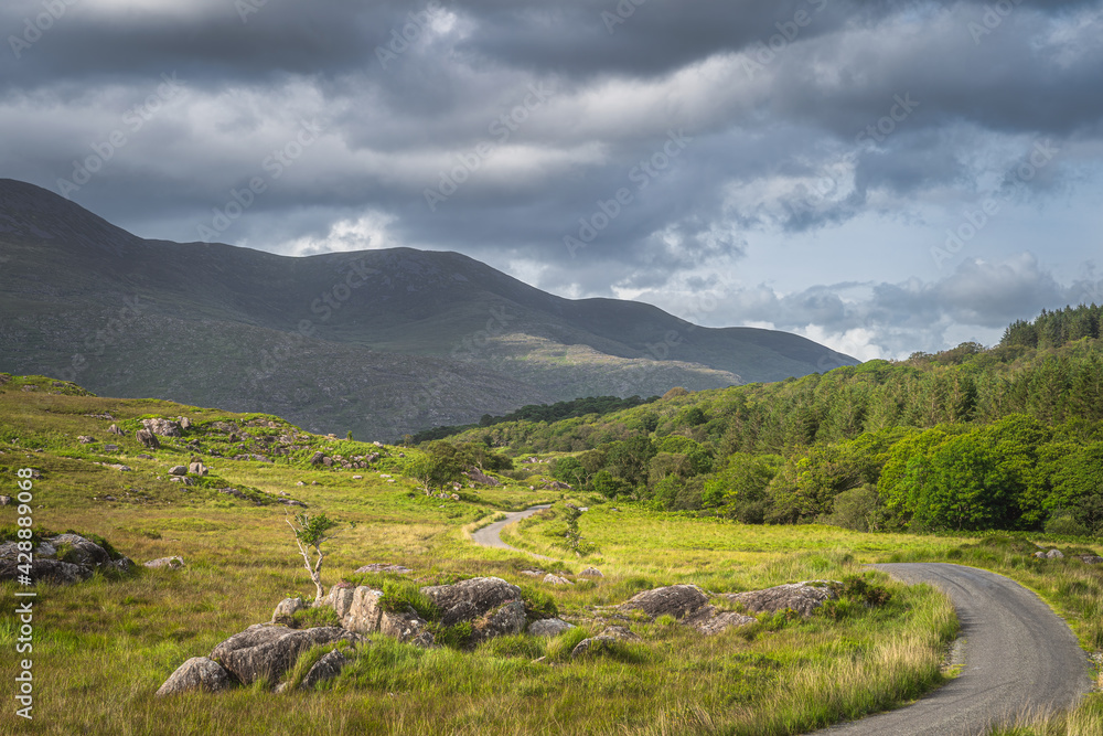 Winding country road leading trough valley, a rough landscape with rocky hills of Molls Gap in MacGillycuddys Reeks mountains, Ring of Kerry, Ireland Winding country road leading trough valley, a rough landscape with rocky hills of Molls Gap in MacGillycuddys Reeks mountains, Ring of Kerry, Ireland